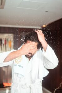 A man in a robe dries his hair in a chic hotel bathroom, splashing water droplets.