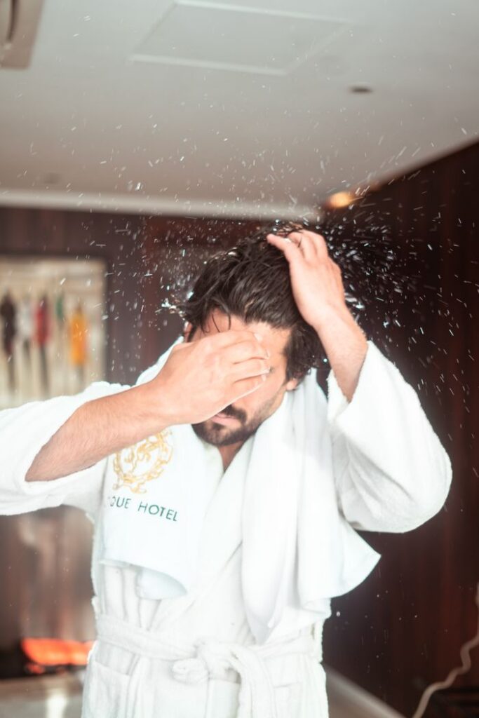 A man in a robe dries his hair in a chic hotel bathroom, splashing water droplets.