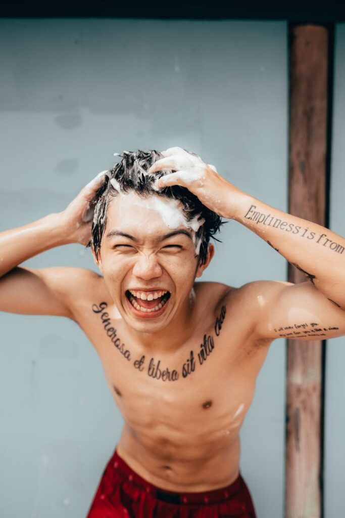 Shirtless man enjoying hair wash with a joyful expression outdoors, showcasing tattoos.