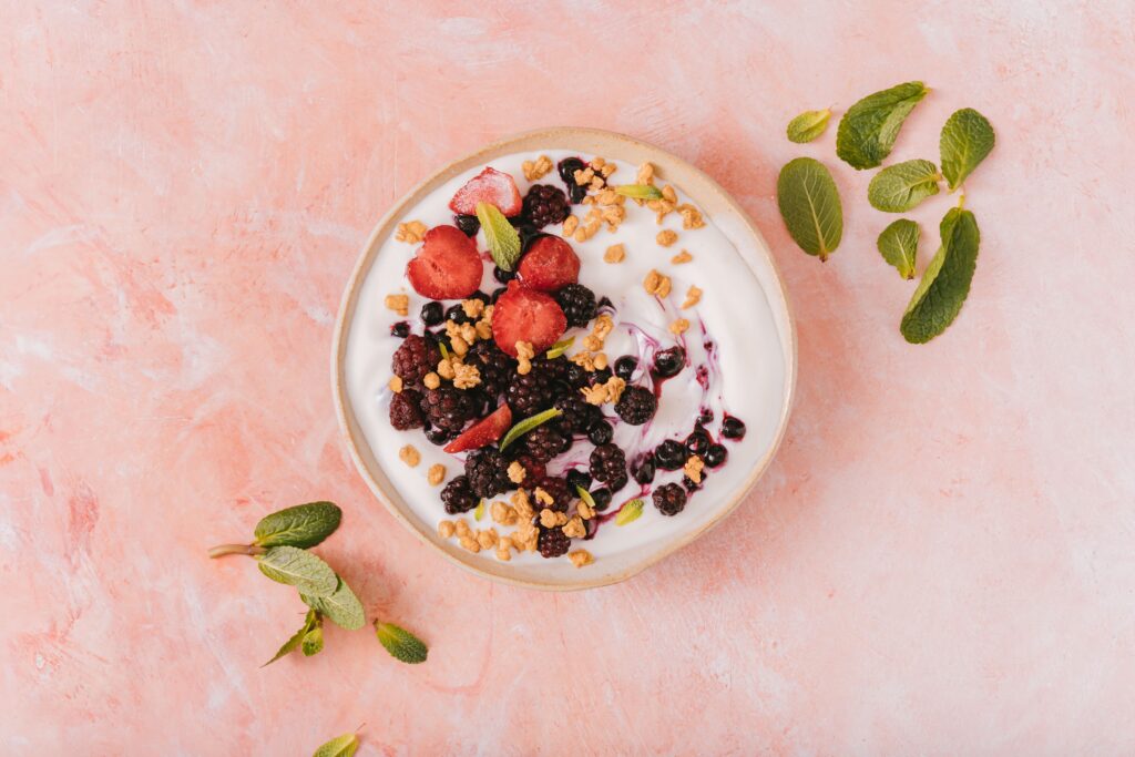 pexels-photo-6823339-6823339 Top view of a vibrant yogurt bowl with mixed berries, granola, and mint leaves on a pink background.