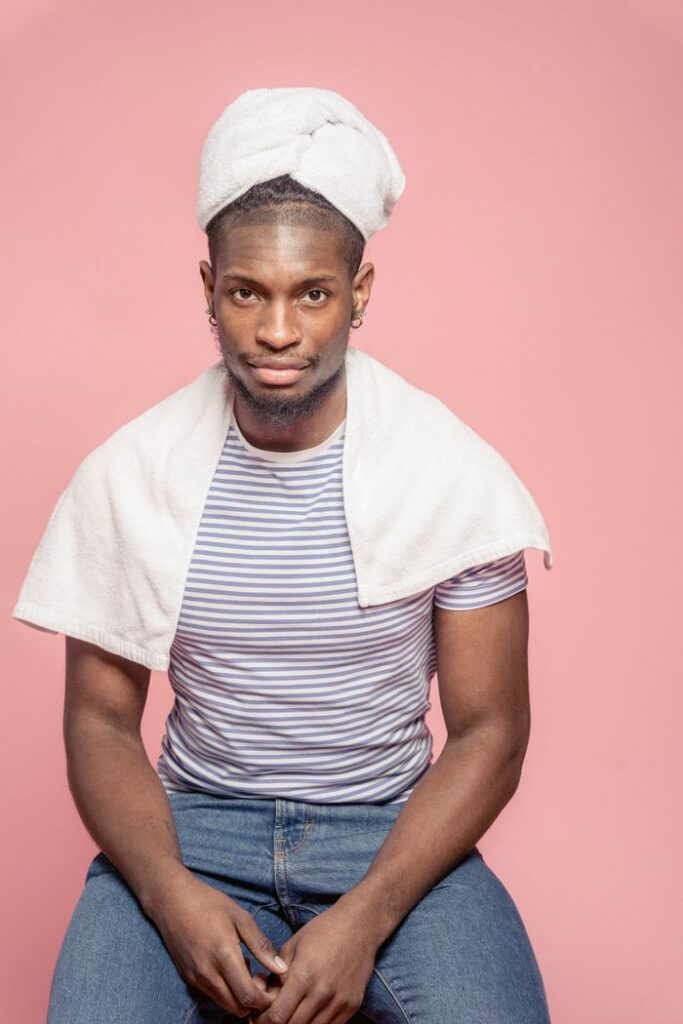 Unshaven African American male client in casual outfit dying hair with towel turban after dying hair in studio against pink background