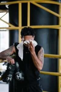 Boxer with tattoos resting in gym, wiping sweat with towel after training session.