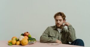 A man in casual attire sits at a table with various vegetables, evoking a thoughtful mood.
