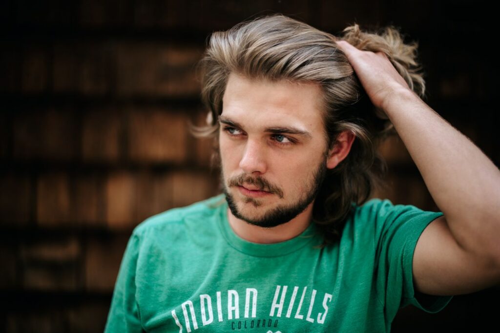 pexels-photo-8786708-8786708 Casual portrait of a man with long brown hair wearing a green t-shirt, hand on hair.