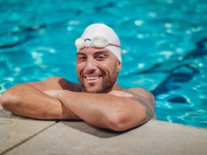 A male swimmer smiles while resting at the edge of a swimming pool, wearing swim cap and goggles.
