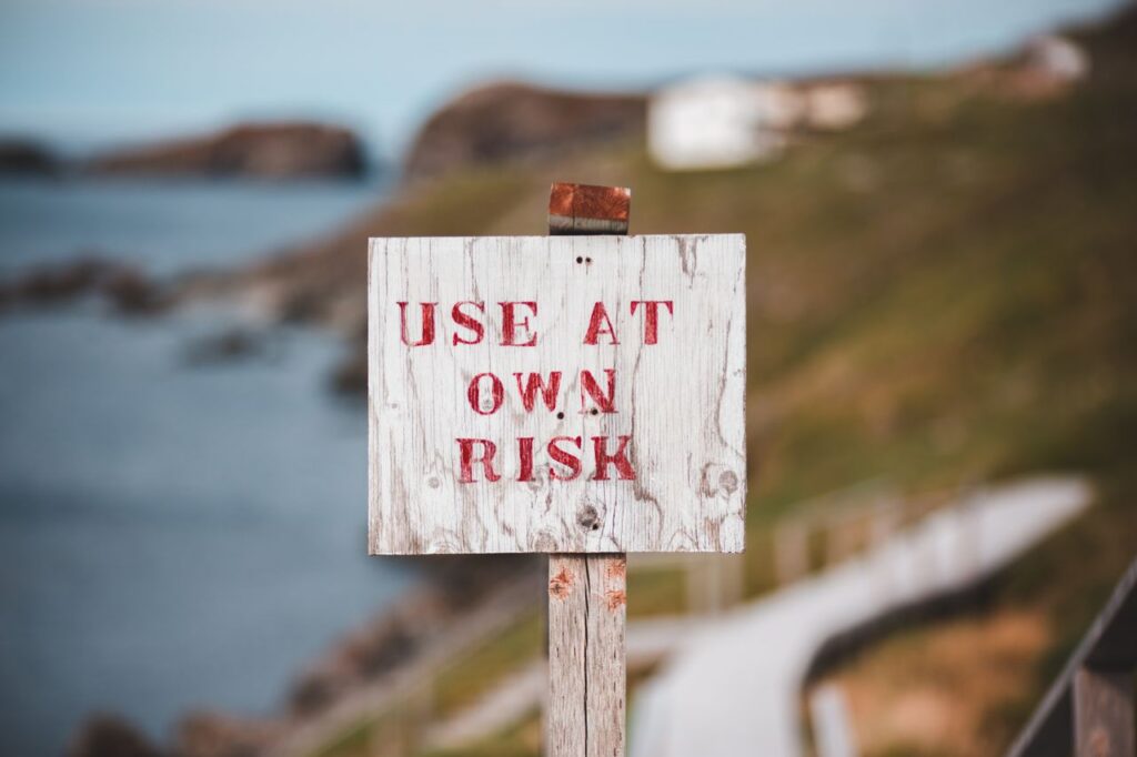 Weathered poled sign with inscription Use At Own Risk located on stiff cliff slope near staircase against blurred sea